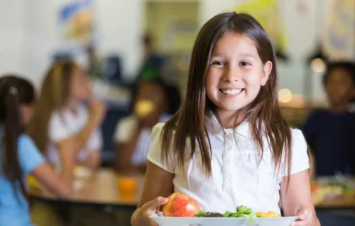 A young girl smiles while holding her lunch tray
